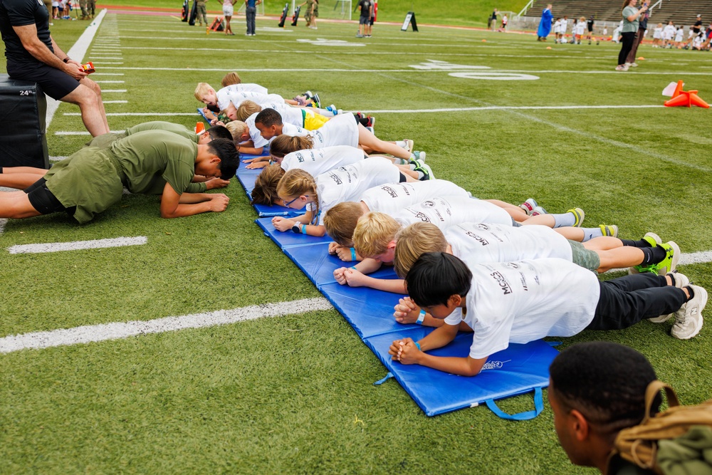 A group of children in white t-shirts are performing a plank exercise on a blue gymnastics mat on a football field. They appear to be participating in a youth fitness or sports camp, guided by adults in military-style olive green uniforms. The event takes place on a marked turf field with additional participants and spectators visible in the background.