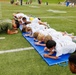 A group of children in white t-shirts are performing a plank exercise on a blue gymnastics mat on a football field. They appear to be participating in a youth fitness or sports camp, guided by adults in military-style olive green uniforms. The event takes place on a marked turf field with additional participants and spectators visible in the background.