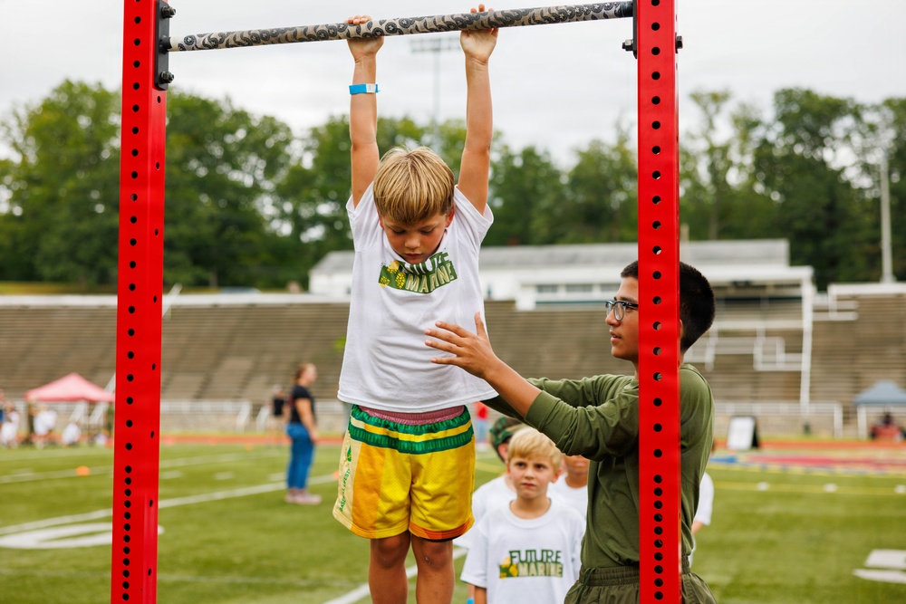 A young blonde boy wearing a white 'Future Marine' t-shirt and yellow shorts hangs from a pull-up bar mounted on a red metal frame, while an older youth in an olive green uniform steadies him with a supportive hand. The activity appears to be part of a youth fitness or military-themed camp event taking place on an athletic field. Other children in matching 'Future Marine' shirts are visible waiting in the background.