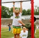 A young blonde boy wearing a white 'Future Marine' t-shirt and yellow shorts hangs from a pull-up bar mounted on a red metal frame, while an older youth in an olive green uniform steadies him with a supportive hand. The activity appears to be part of a youth fitness or military-themed camp event taking place on an athletic field. Other children in matching 'Future Marine' shirts are visible waiting in the background.