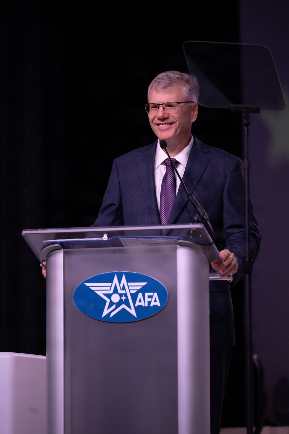 A middle-aged man with gray hair and glasses is speaking at a podium during a formal event. The podium displays the AFA (Air Force Association) logo, indicating this is an official AFA function or conference. The speaker is smiling and appears engaged with the audience.