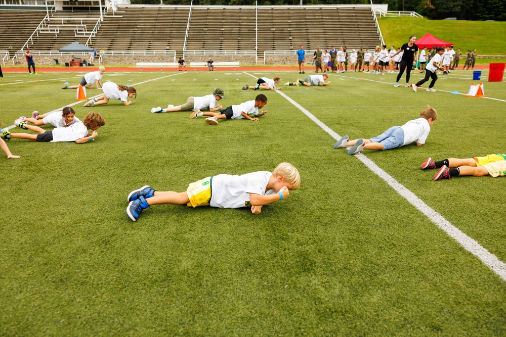 A group of children are participating in an organized physical fitness event on a green turf football field inside a stadium. The children are performing a low crawl or plank-style exercise simultaneously, spread across the field in rows. Adults and supervisors are visible in the background near a red canopy tent, overseeing the activity.