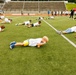 A group of children are participating in an organized physical fitness event on a green turf football field inside a stadium. The children are performing a low crawl or plank-style exercise simultaneously, spread across the field in rows. Adults and supervisors are visible in the background near a red canopy tent, overseeing the activity.