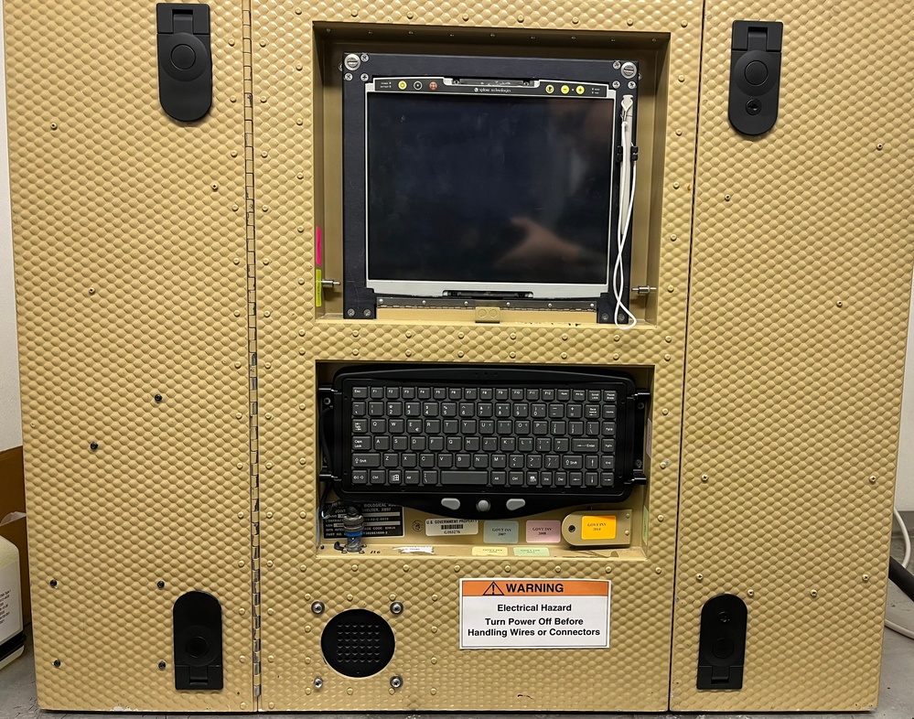 A ruggedized military or government computing workstation housed in a tan/beige armored enclosure with a dimple-pattern surface. The unit features an embedded flat-panel display, a compact keyboard in a recessed bay, and multiple latching mechanisms suggesting it is designed for field or mobile deployment. A warning label indicates electrical hazard precautions are required before handling internal wiring.