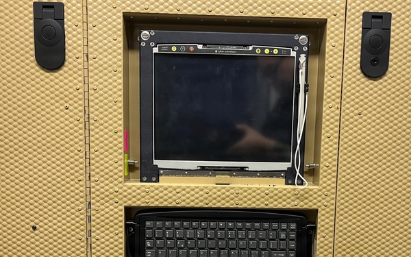 A ruggedized military or government computing workstation housed in a tan/beige armored enclosure with a dimple-pattern surface. The unit features an embedded flat-panel display, a compact keyboard in a recessed bay, and multiple latching mechanisms suggesting it is designed for field or mobile deployment. A warning label indicates electrical hazard precautions are required before handling internal wiring.