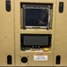 A ruggedized military or government computing workstation housed in a tan/beige armored enclosure with a dimple-pattern surface. The unit features an embedded flat-panel display, a compact keyboard in a recessed bay, and multiple latching mechanisms suggesting it is designed for field or mobile deployment. A warning label indicates electrical hazard precautions are required before handling internal wiring.