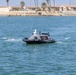 An unmanned surface vessel (USV) is operating on calm coastal waters near a shoreline with residential buildings and palm trees in the background. The dark-colored autonomous boat is equipped with a camera/sensor turret on top and bears the warning text 'KEEP BACK - USV' on its hull. The vessel appears to be underway, creating small wake patterns on both sides.