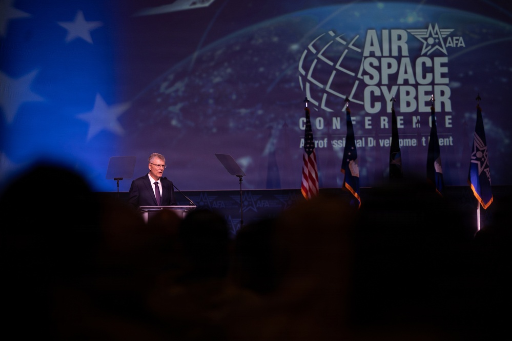 A speaker addresses an audience from a podium at the AFA (Air Force Association) Air Space Cyber Conference, described as a Professional Development Event. The stage is dramatically lit with a large projection screen displaying the conference branding, and multiple flags are visible behind the speaker. The audience is visible in silhouette in the foreground.
