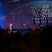 A speaker addresses an audience from a podium at the AFA (Air Force Association) Air Space Cyber Conference, described as a Professional Development Event. The stage is dramatically lit with a large projection screen displaying the conference branding, and multiple flags are visible behind the speaker. The audience is visible in silhouette in the foreground.