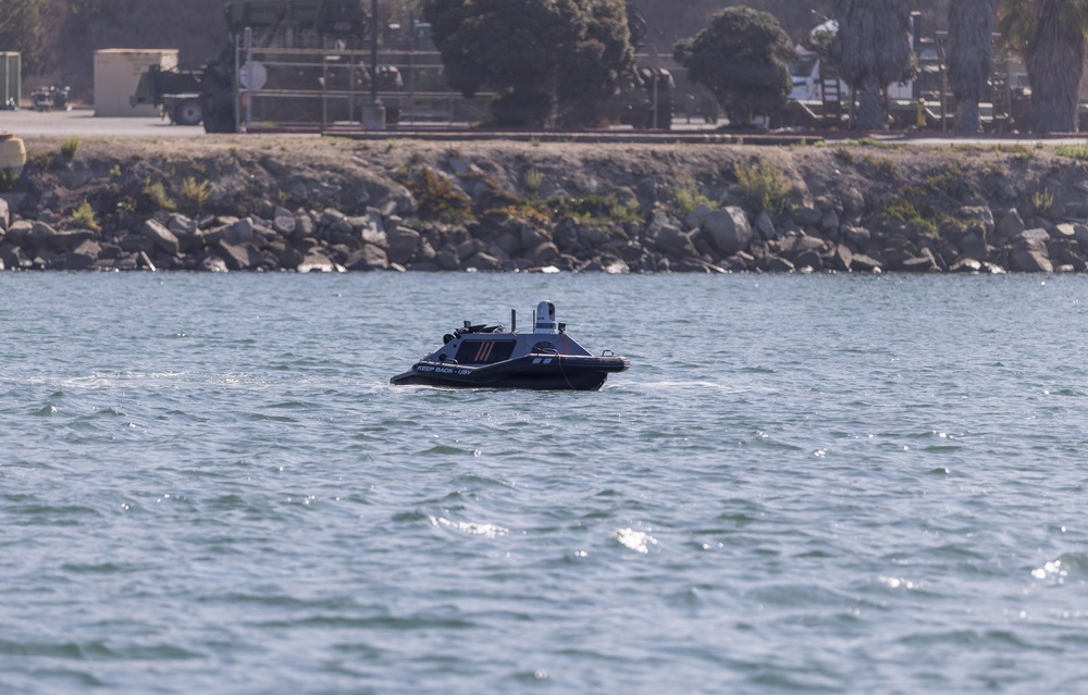 A small, dark-colored unmanned surface vessel (USV) is operating on open water near a rocky shoreline. The vessel bears visible text reading 'KEEP BACK - USV' on its hull, indicating it is an autonomous or remotely operated watercraft. A military or industrial facility with vehicles and structures is visible in the background beyond the rock breakwater.
