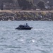 A small, dark-colored unmanned surface vessel (USV) is operating on open water near a rocky shoreline. The vessel bears visible text reading 'KEEP BACK - USV' on its hull, indicating it is an autonomous or remotely operated watercraft. A military or industrial facility with vehicles and structures is visible in the background beyond the rock breakwater.