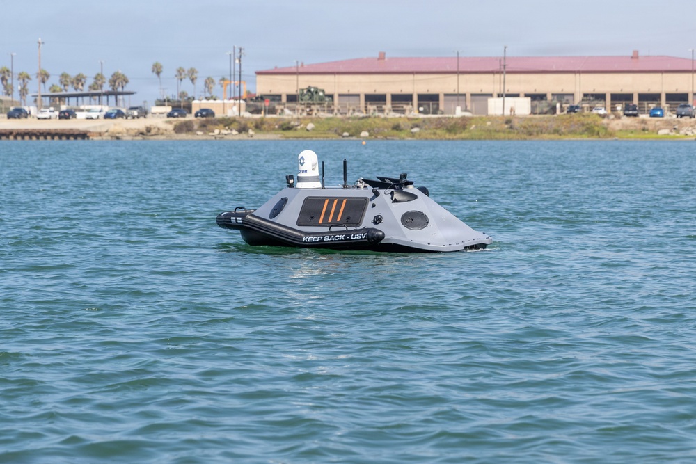 The image shows an unmanned surface vessel (USV) floating on calm harbor waters. The gray autonomous watercraft is marked 'KEEP BACK - USV' and is equipped with sensors, cameras, and communication antennas on top. A large industrial or military facility with palm trees is visible in the background along the shoreline.