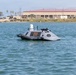 The image shows an unmanned surface vessel (USV) floating on calm harbor waters. The gray autonomous watercraft is marked 'KEEP BACK - USV' and is equipped with sensors, cameras, and communication antennas on top. A large industrial or military facility with palm trees is visible in the background along the shoreline.