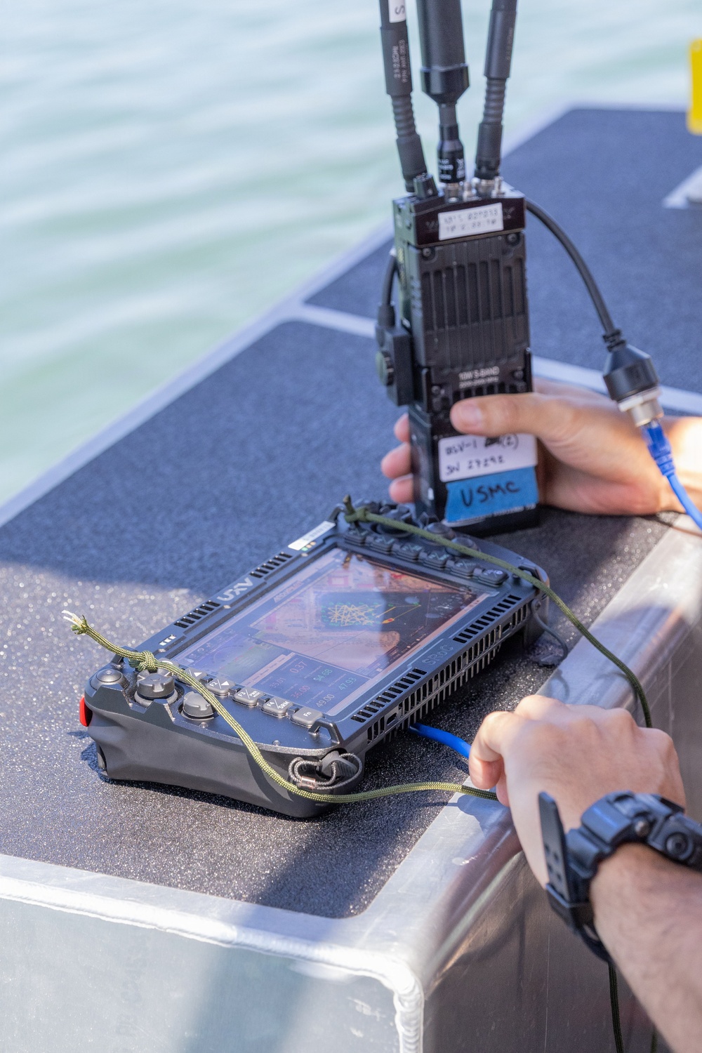 A person is handling military-grade communications and UAV control equipment on what appears to be a watercraft. The equipment includes a rugged tablet-style UAV ground control station and a radio transceiver unit labeled 'USMC', connected via blue Ethernet and other cables. The setting appears to be an outdoor maritime environment in daylight.