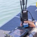 A person is handling military-grade communications and UAV control equipment on what appears to be a watercraft. The equipment includes a rugged tablet-style UAV ground control station and a radio transceiver unit labeled 'USMC', connected via blue Ethernet and other cables. The setting appears to be an outdoor maritime environment in daylight.