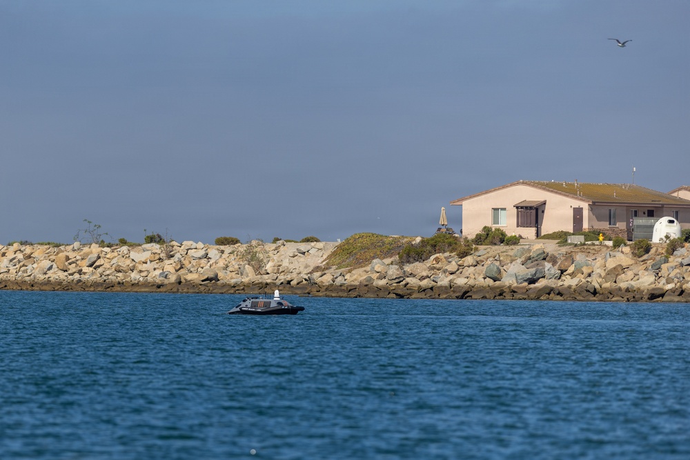 A coastal waterway scene featuring a small dark-colored vessel on calm blue water in the foreground. A rocky breakwater or jetty runs along the background, with a residential building visible behind it. The sky is overcast with a hazy blue-gray tone, and a single bird is visible in flight near the upper right.