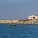 A coastal waterway scene featuring a small dark-colored vessel on calm blue water in the foreground. A rocky breakwater or jetty runs along the background, with a residential building visible behind it. The sky is overcast with a hazy blue-gray tone, and a single bird is visible in flight near the upper right.