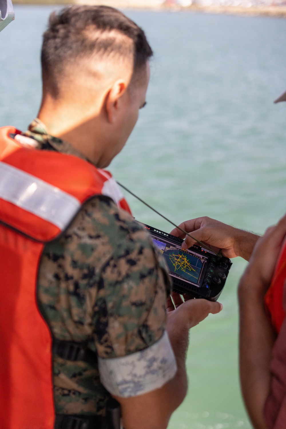 A military personnel wearing USMC MARPAT camouflage uniform and an orange life vest is examining a handheld electronic device with a screen displaying what appears to be a navigation or sonar/mapping interface with yellow grid lines. The setting appears to be near or on a body of water, likely during a maritime training exercise. A second person partially visible on the right is also holding or pointing at the device.