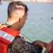 A military personnel wearing USMC MARPAT camouflage uniform and an orange life vest is examining a handheld electronic device with a screen displaying what appears to be a navigation or sonar/mapping interface with yellow grid lines. The setting appears to be near or on a body of water, likely during a maritime training exercise. A second person partially visible on the right is also holding or pointing at the device.
