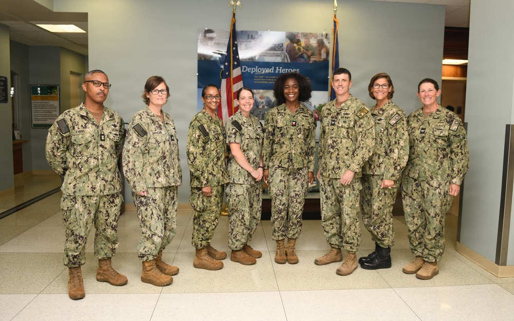 Eight U.S. Navy personnel in Navy Working Uniform (NWU Type III) camouflage are posed for a group photo in what appears to be a military facility lobby. They are standing in front of a 'Deployed Heroes' banner and U.S. flags. The group consists of both male and female service members of varying ranks.