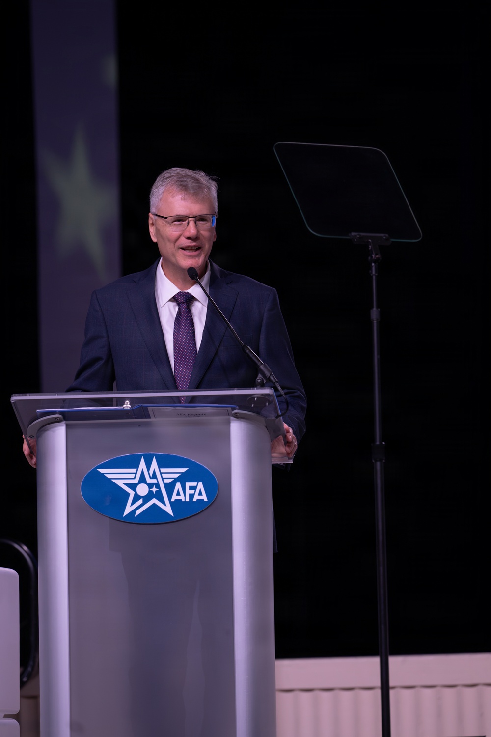 A man in a dark navy suit with a purple tie is speaking at a podium bearing the AFA (Air Force Association) logo. He appears to be delivering a formal address or presentation at what looks like an official AFA event or conference. A teleprompter screen is visible to his right on a stand.