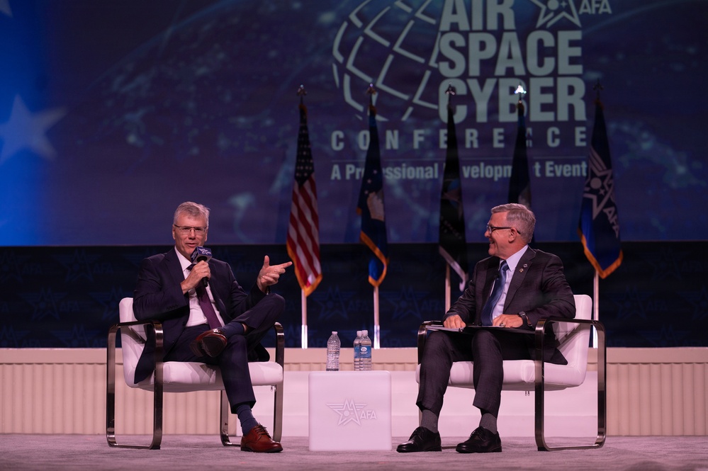 Two men in business suits are seated on a stage at the AFA (Air Force Association) Air, Space &amp; Cyber Conference, a professional development event. The man on the left is speaking into a microphone while gesturing, and the man on the right is listening and smiling while holding a folder or notepad. Multiple flags, including the U.S. flag, are displayed on stage behind them.