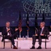 Two men in business suits are seated on a stage at the AFA (Air Force Association) Air, Space &amp; Cyber Conference, a professional development event. The man on the left is speaking into a microphone while gesturing, and the man on the right is listening and smiling while holding a folder or notepad. Multiple flags, including the U.S. flag, are displayed on stage behind them.