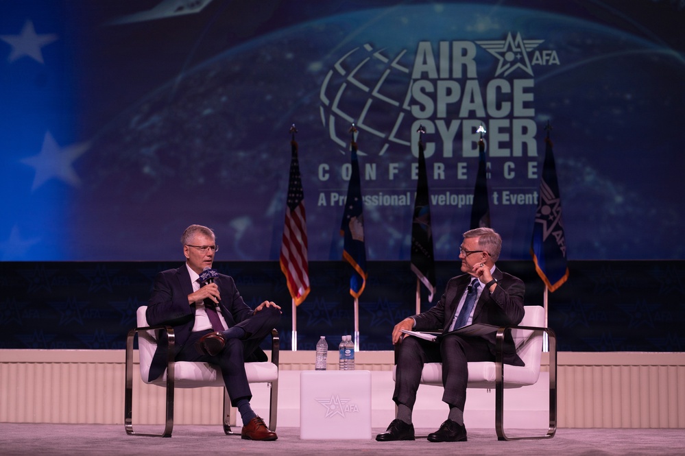 Two men in business attire are seated on a stage at the AFA (Air Force Association) Air Space Cyber Conference, a professional development event. The man on the left is speaking into a microphone while the man on the right listens attentively with his hand raised to his chin. The backdrop displays the conference branding along with multiple flags including the American flag.