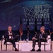 Two men in business attire are seated on a stage at the AFA (Air Force Association) Air Space Cyber Conference, a professional development event. The man on the left is speaking into a microphone while the man on the right listens attentively with his hand raised to his chin. The backdrop displays the conference branding along with multiple flags including the American flag.