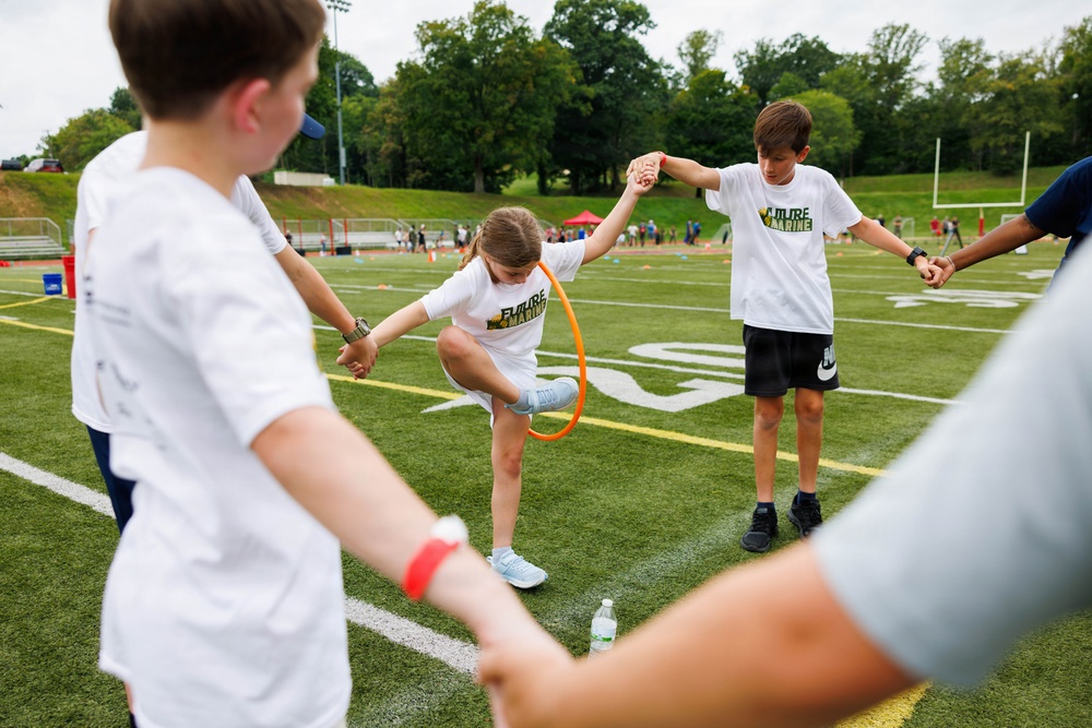 A group of children participate in an outdoor team-building activity on a football field, passing an orange hula hoop along a chain of held hands without breaking their grip. The children are wearing matching white 'Future Marine' t-shirts, suggesting an organized youth program or camp event. The activity takes place on a well-maintained artificial turf field with bleachers and trees visible in the background.