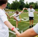 A group of children participate in an outdoor team-building activity on a football field, passing an orange hula hoop along a chain of held hands without breaking their grip. The children are wearing matching white 'Future Marine' t-shirts, suggesting an organized youth program or camp event. The activity takes place on a well-maintained artificial turf field with bleachers and trees visible in the background.