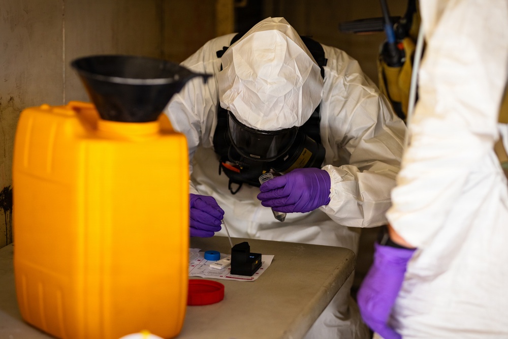 Two individuals wearing full hazmat/PPE suits with respirator masks and purple nitrile gloves are conducting what appears to be a field chemical or biological sample analysis at a workbench. The central figure is closely examining a small sample container while manipulating a swab or testing instrument over a test kit layout. The setting appears to be an enclosed or semi-enclosed operational environment with hazardous material containers nearby.
