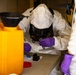 Two individuals wearing full hazmat/PPE suits with respirator masks and purple nitrile gloves are conducting what appears to be a field chemical or biological sample analysis at a workbench. The central figure is closely examining a small sample container while manipulating a swab or testing instrument over a test kit layout. The setting appears to be an enclosed or semi-enclosed operational environment with hazardous material containers nearby.