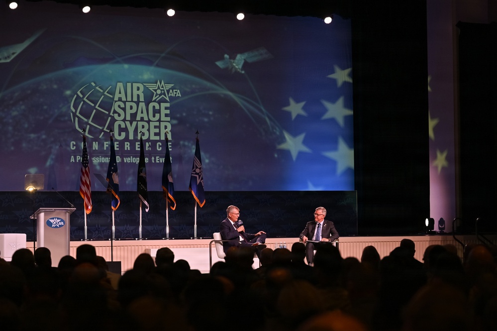 Two individuals are seated on stage at the AFA (Air Force Association) Air Space Cyber Conference, a professional development event. One person is holding a microphone, suggesting a moderated discussion or interview format. A large audience is visible in the foreground, and multiple flags are displayed on stage alongside a podium.