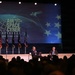 Two individuals are seated on stage at the AFA (Air Force Association) Air Space Cyber Conference, a professional development event. One person is holding a microphone, suggesting a moderated discussion or interview format. A large audience is visible in the foreground, and multiple flags are displayed on stage alongside a podium.