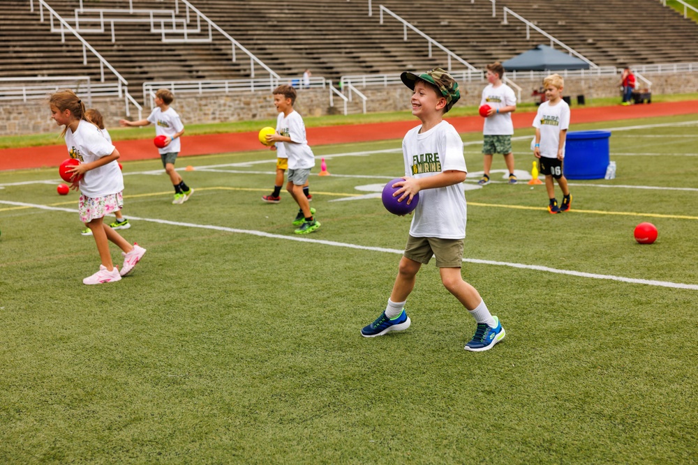 A group of young children participate in an outdoor sports camp or clinic on a turf athletic field. The children are wearing matching white t-shirts with 'Future Marine' printed on them and are carrying various colored dodgeballs. The activity appears to be an organized drill or game involving ball-handling on a stadium field with bleachers visible in the background.