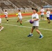A group of young children participate in an outdoor sports camp or clinic on a turf athletic field. The children are wearing matching white t-shirts with 'Future Marine' printed on them and are carrying various colored dodgeballs. The activity appears to be an organized drill or game involving ball-handling on a stadium field with bleachers visible in the background.