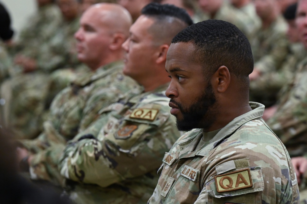 A group of military personnel in OCP (Operational Camouflage Pattern) uniforms are seated in formation, likely attending a briefing, ceremony, or formal gathering. The foreground subject is a bearded male service member wearing a 'QA' (Quality Assurance) patch and a 'U.S. Air Force' tape on his uniform. Multiple rows of similarly uniformed personnel are visible in the background, slightly out of focus.
