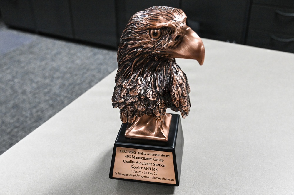 The image shows a bronze/copper-colored eagle head trophy mounted on a black base with an engraved plaque. The award is the AFRC MXG Quality Assurance Award presented to the 403 Maintenance Group Quality Assurance Section at Keesler AFB, Mississippi, for the period 1 Jan 25 - 31 Dec 25. The trophy is placed on a light gray surface, likely a table or desk.