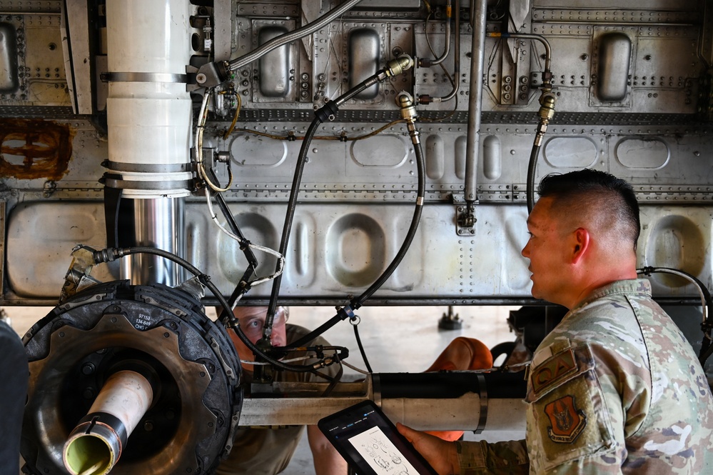 Two military personnel in OCP (Operational Camouflage Pattern) uniforms are performing maintenance on what appears to be a large military aircraft's landing gear assembly. One airman is consulting a tablet device displaying technical diagrams while the other inspects the undercarriage components. The image shows the underside of the aircraft with visible hydraulic lines, structural panels, and a nose or main landing gear strut with tire.