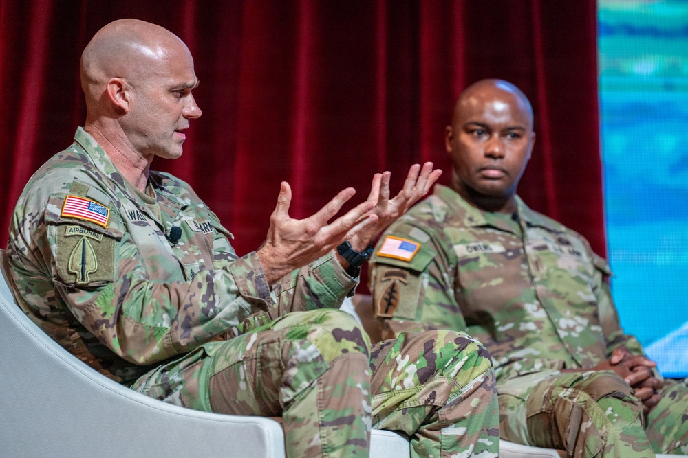 Two U.S. Army soldiers in Operational Camouflage Pattern (OCP) uniforms are seated on a stage, apparently participating in a panel discussion or conference. The soldier on the left is actively speaking and gesturing with both hands, while the soldier on the right listens attentively. A red curtain serves as the backdrop, suggesting a formal event or symposium setting.
