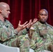 Two U.S. Army soldiers in Operational Camouflage Pattern (OCP) uniforms are seated on a stage, apparently participating in a panel discussion or conference. The soldier on the left is actively speaking and gesturing with both hands, while the soldier on the right listens attentively. A red curtain serves as the backdrop, suggesting a formal event or symposium setting.