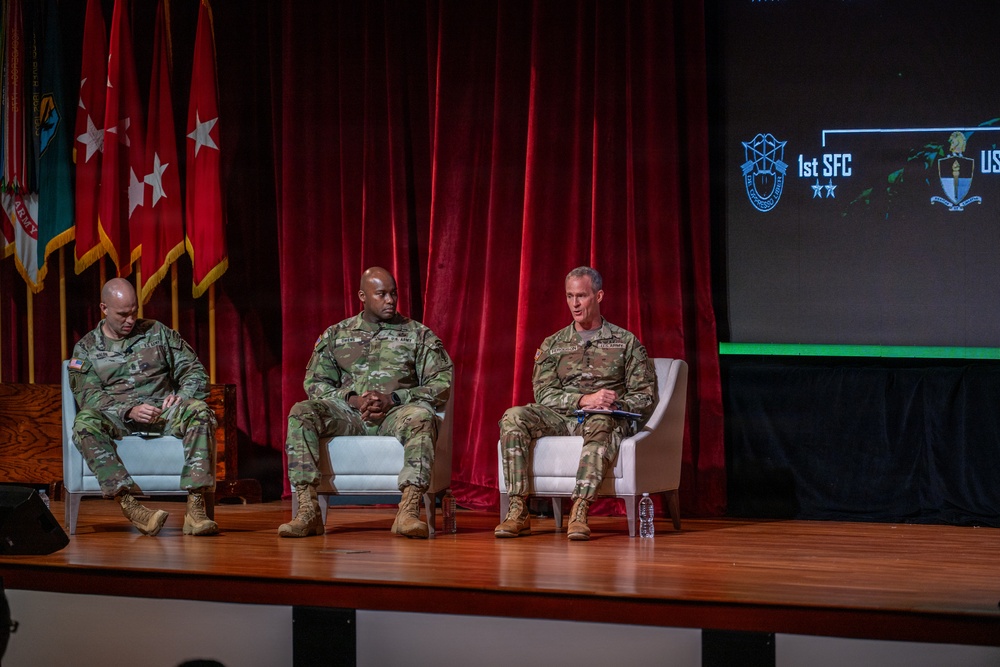 Three U.S. Army soldiers in OCP (Operational Camouflage Pattern) uniforms are seated in white chairs on a wooden stage during what appears to be a panel discussion or briefing. A projection screen to the right displays '1st SFC' along with military unit insignia. Military flags and a red curtain backdrop are visible behind them.