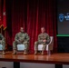 Three U.S. Army soldiers in OCP (Operational Camouflage Pattern) uniforms are seated in white chairs on a wooden stage during what appears to be a panel discussion or briefing. A projection screen to the right displays '1st SFC' along with military unit insignia. Military flags and a red curtain backdrop are visible behind them.