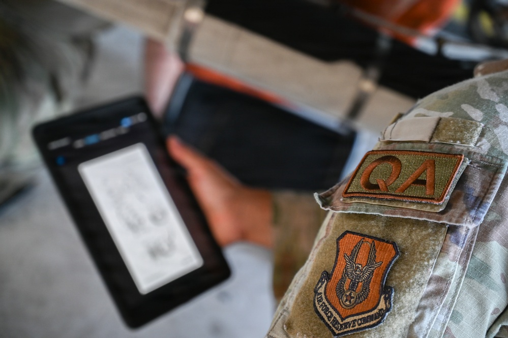 A close-up image of a military personnel wearing an OCP (Operational Camouflage Pattern) uniform with visible patches, holding a tablet device. The uniform displays a 'QA' (Quality Assurance) tab patch and an Air Force Reserve Command shield patch on the sleeve. The focus is on the uniform patches while the tablet in the background is intentionally blurred.