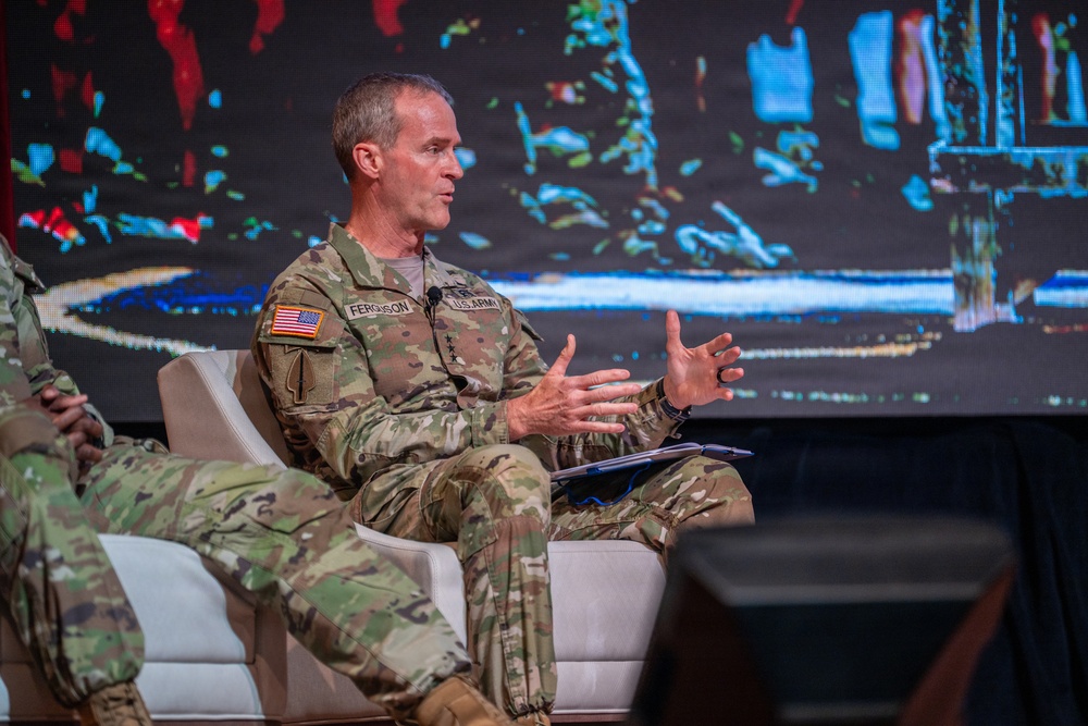 A U.S. Army officer identified by name tape as 'Ferguson' is seated on a white chair on a stage, appearing to speak at a panel or conference event. He is wearing OCP (Operational Camouflage Pattern) uniform with U.S. Army insignia, an American flag patch, and what appears to be general officer rank insignia. A large digital display screen is visible in the background.