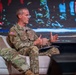 A U.S. Army officer identified by name tape as 'Ferguson' is seated on a white chair on a stage, appearing to speak at a panel or conference event. He is wearing OCP (Operational Camouflage Pattern) uniform with U.S. Army insignia, an American flag patch, and what appears to be general officer rank insignia. A large digital display screen is visible in the background.