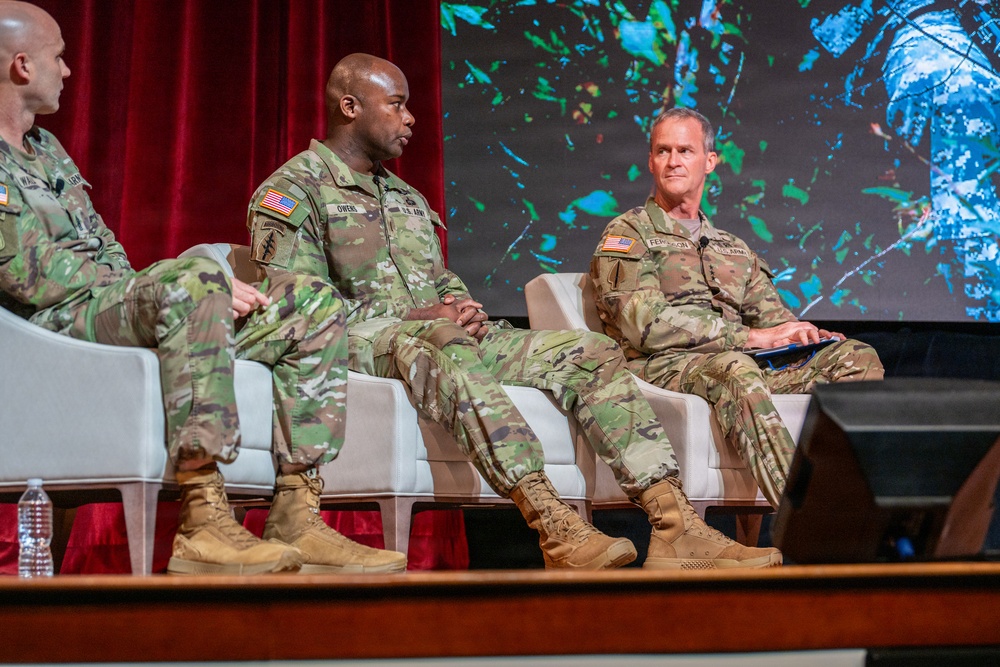 Three U.S. Army soldiers in OCP (Operational Camouflage Pattern) uniforms are seated on a stage panel, appearing to participate in a formal military symposium or conference. The central figure, identified by name tape as 'OWENS,' is speaking while the soldier to the right, identified as 'FERGUSON,' listens and holds a tablet or folder. A large display screen with a nature-themed background is visible behind them.