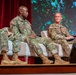 Three U.S. Army soldiers in OCP (Operational Camouflage Pattern) uniforms are seated on a stage panel, appearing to participate in a formal military symposium or conference. The central figure, identified by name tape as 'OWENS,' is speaking while the soldier to the right, identified as 'FERGUSON,' listens and holds a tablet or folder. A large display screen with a nature-themed background is visible behind them.