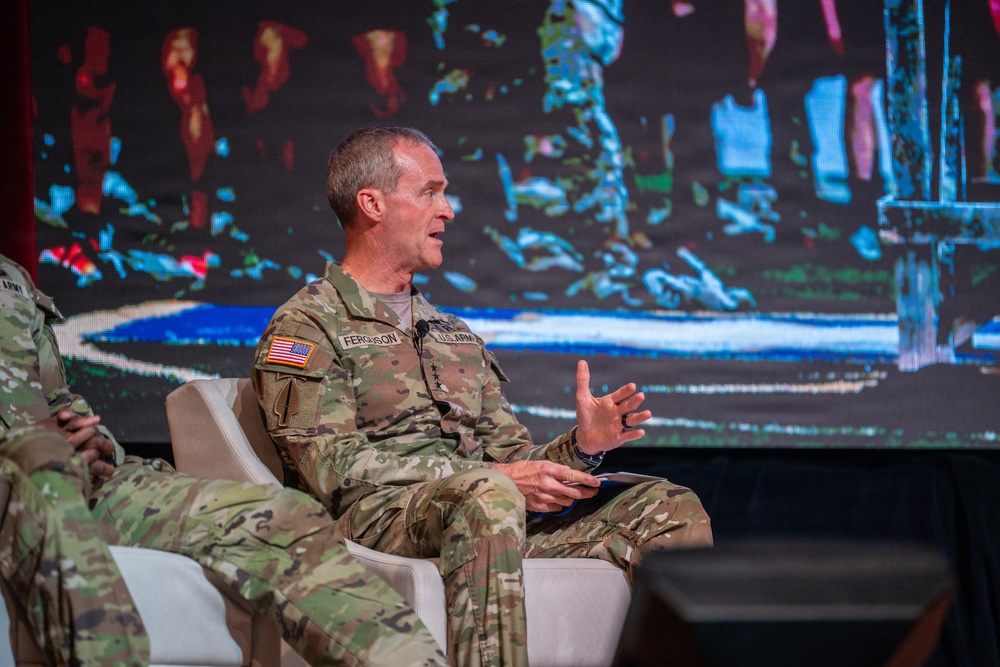 A U.S. Army soldier identified by name tape as 'Ferguson' is seated on a white chair on a stage, actively speaking and gesturing with his hands during what appears to be a panel discussion or conference. He is wearing the Operational Camouflage Pattern (OCP) uniform with a U.S. flag patch and 'U.S. ARMY' tape visible. Another soldier in the same uniform is partially visible on the left side of the frame.